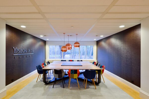 Vergaderruimte in Ypsilon Park Lounge met een grote houten tafel, kleurrijke stoelen en drie koperen hanglampen.